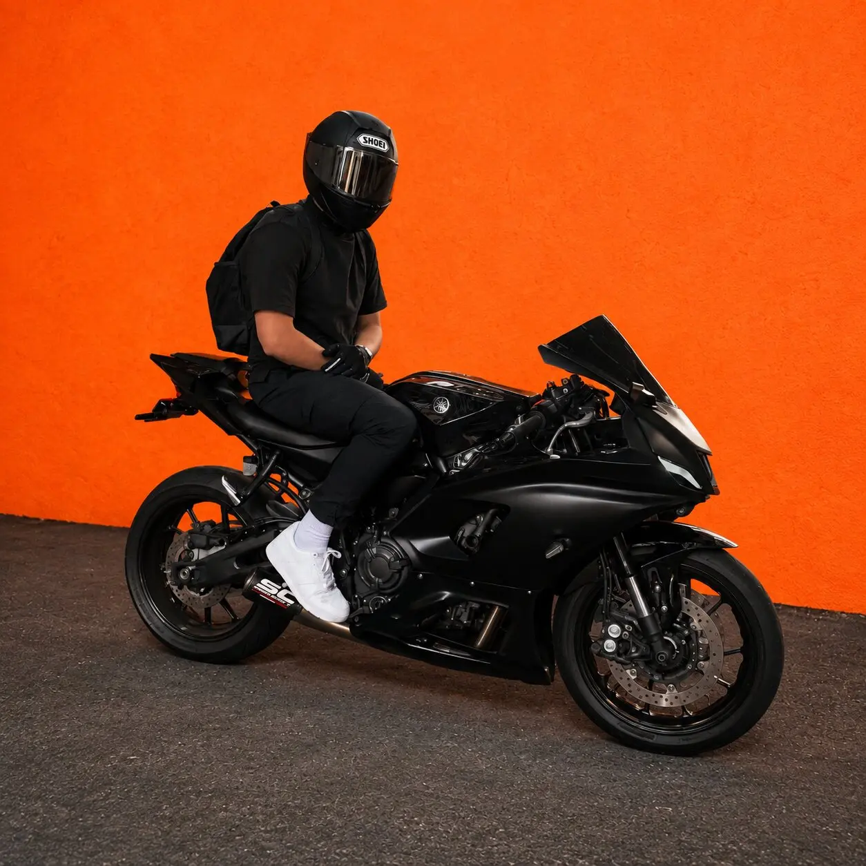 Bryn seated on a black motorcycle in front of an orange wall